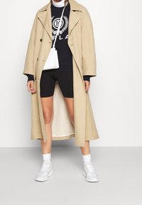 Beige trenchcoat med breda slag, lager på en svart sweatshirt och svarta cykelshorts. Vita sneakers och en liten vit axelväska.