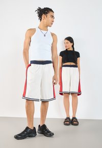 Shorts de sport blancs avec des panneaux latéraux rouges et des accents bleu marine, portés avec un débardeur blanc et un crop top noir ; baskets noires et tongs.