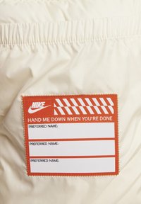 Chaqueta acolchada blanca con una etiqueta naranja que tiene texto blanco que dice "PASÁMELA CUANDO TERMINES" y secciones para nombres.