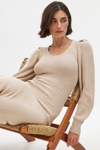Beige ribbad klänning med långa ärmar och subtila axeldetaljer. Modell som sitter på en trästol med vävd sits.