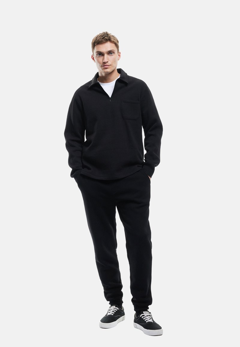 Sort pullover med lynlås krave og brystlomme, parret med matchende sorte bukser. Modellen bærer en hvid t-shirt og sorte sneakers.