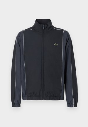 Veste zippée noire avec un col montant, des bandes latérales blanches, des poignets  élastiques et un petit logo vert sur la poitrine gauche. Texture de tissu lisse.