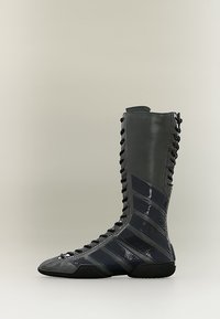 Ψηλό μαύρο μποτάκι με κορδόνια, γυαλιστερές διαγώνιες ρίγες και φερμουάρ στο πλάι, εκτεθειμένο σε απλό ανοιχτό φόντο.