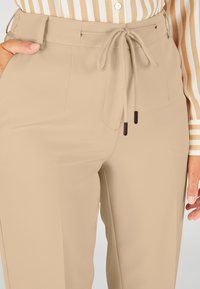 Beige, hooggetailleerde broek van gladde stof met een trekkoord aan de taille, voorzien van twee zijzakken en subtiele stikseldetails.