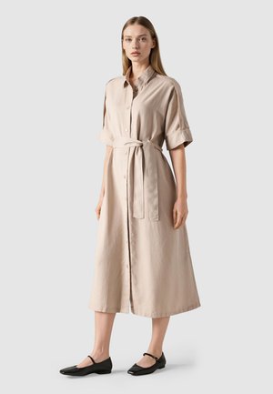 Žena stojící v béžových midi šatech zapnutých na knoflíky, s vyhrnutými rukávy a páskem v pase, v černých balerínách Mary Jane.