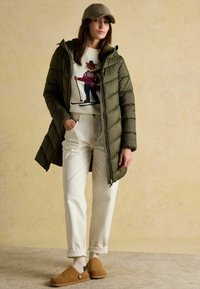 Olivovo zelená prešívaná bunda, biela sveter s grafikou lyžiara, svetlo béžové nohavice a hnedé semišov�é dreváky so zlatým detailom na gombíku.