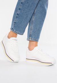 Personne portant des baskets compensées blanches avec des rayures pastel et un jean bleu raccourci, debout sur un sol blanc.