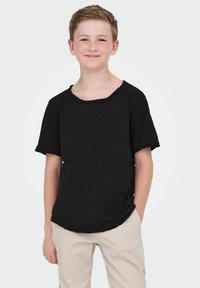 Czarna koszulka z krótkim rękawem o szerokim dekolcie i surowych brzegach, noszona z beżowymi spodniami. Materiał wydaje się miękki i casualowy.