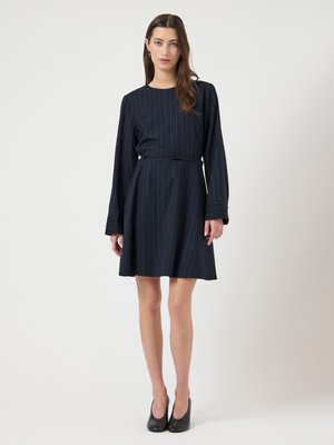 Donna che indossa un vestito a righe blu navy con cintura, maniche lunghe e tacchi chiusi neri, in piedi contro uno sfondo bianco semplice.
