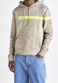 Beige hoodie med tonala geometriska mönster, neongrön accentrand, justerbara dragsnören och en framficka i kengurustil.