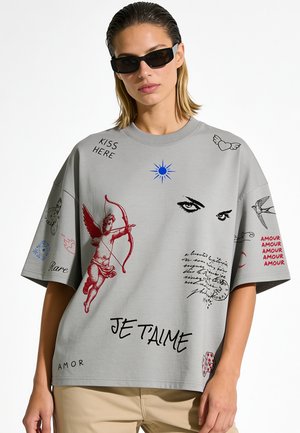 Persona che indossa una t-shirt grigia oversize con Cupido, occhi, testo "Je t'aime," "Kiss here," e disegni grafici rossi, neri e blu, abbinata a pantaloni beige.