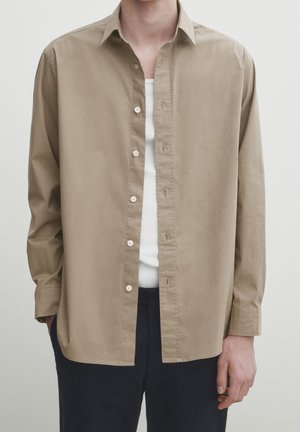 Mand iført en åben beige langærmet skjorte over en hvid ribbet tanktop med sorte bukser mod en ensfarvet baggrund.