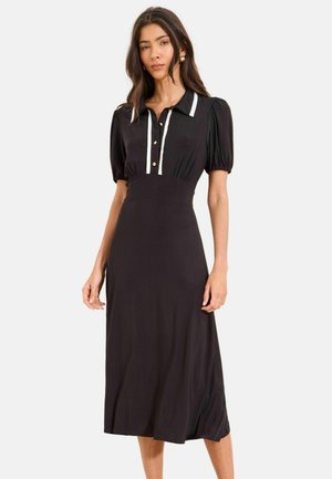 Kobieta ubrana w czarną midi sukienkę z bufkami, białym kołnierzykiem i plisą na guziki, stojąca na jednolitym tle.