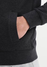 Felpa grigia scura con tessuto strutturato, polsini a costine e tasca frontale. Una mano è inserita nella tasca. Design semplice e stile informale.