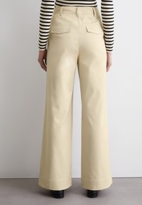 Wijde pantalons van een lichtbeige stof, voorzien van twee achterzakken en een gladde textuur, gedragen met zwarte platformlaarzen.