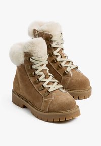 Beige Wildleder-Ankle-Boots mit Kunstfellkragen, dicker beiger Gummisohle und beigen Schnürsenkeln, ausgestattet mit Metallösen und Nähakzenten.