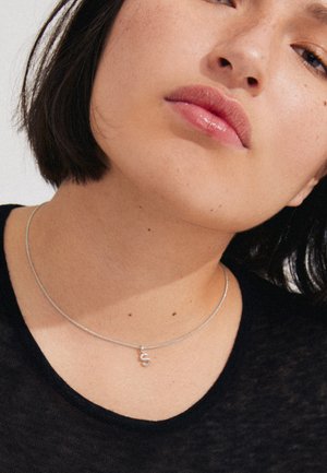 Silberne Kettenhalskette mit einem "S"-Anhänger, getragen auf einem nackten Hals gegen ein schwarzes Shirt. Glatte Textur und schlichtes Design.