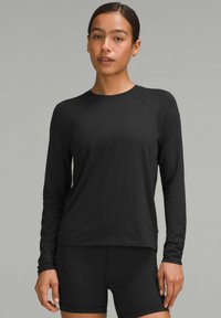 Crna majica dugih rukava za sport s okruglim izrezom i raglan rukavima, izrađena od glatkog, rastezljivog materijala. Vidljive crne kratke hlače.