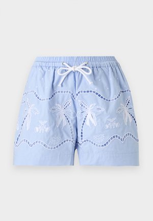 Femme portant une chemise oversize rayée légère, un débardeur blanc, un short brodé bleu clair et des sandales marron, debout devant un fond uni.