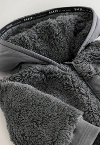 Sivá bunda s kapucňou z mäkkej, chlpaté textúrovanej tkaniny s hladkým vonkajším povrchom. Značka je umiestnená na vnútornej podšívke.