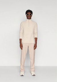 Beige vest met ritssluiting, lichtbeige broek, witte sneakers en oversized zonnebril. Eenvoudig ontwerp met gestructureerde stof en relaxte pasvorm.