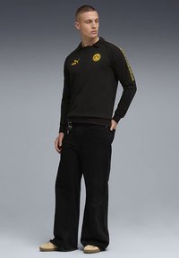 Crni pulover s dugim rukavima i žutim logotipom, s ovratnikom i prugastim detaljima na rukavima, uparen s širokim crnim hlačama i bež cipelama.