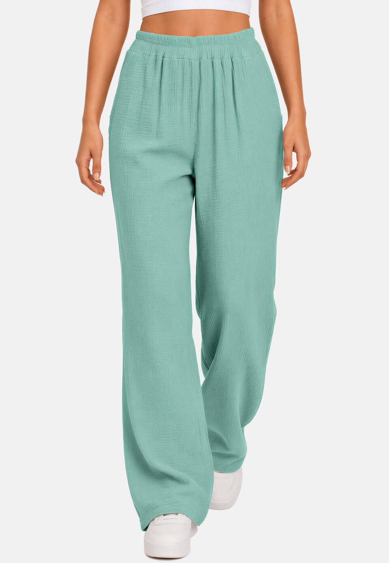 Femme portant un pantalon ample texturé vert menthe et des baskets blanches, montrée de la taille aux pieds sur fond blanc.