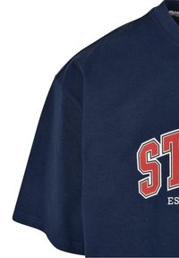 Námořnická modrá bavlněná tričko s krátkým rukávem. Na přední straně má červený a bílý potisk loga "ST". Hladká textura, standardní kulatý výstřih.