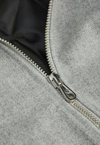 Sivi zip-up odjevni predmet koji prikazuje mekan, teksturiran materijal s srebrnim zatvaračem i kontrastnom crnom podlogom. Prikaz detalja zatvarača izbliza.