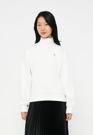 Kvinna med långt svart hår iförd en vit zip-up sweatshirt och svart plisserad kjol, stående mot en enkel vit bakgrund.