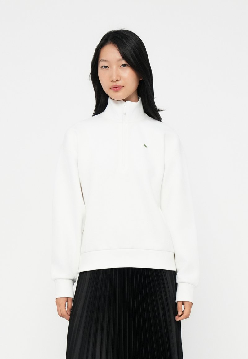 Donna con lunghi capelli neri, che indossa una felpa bianca con zip e una gonna plissettata nera, in piedi contro uno sfondo bianco semplice.