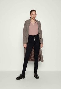 Nő hosszú kockás kabátban, mályva színű galléros pulóverben, magas derekú fekete skinny farmernadrágban és fekete fűzős cipőben áll egy fehér falnak támaszkodva.