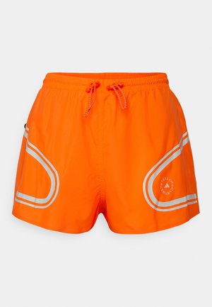 Shorts de sport orange vif avec des bandes réfléchissantes argentées, une taille élastique et des cordons de serrage, présentant un logo circulaire sur la jambe droite.