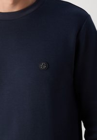 Námorně modrá mikina vyrobená z hladkého materiálu, s kulatou černou nášivkou na hrudi a jemnými detaily prošívání.