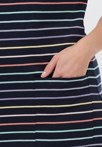 Nawojna sukienka w poziome paski w pastelowych kolorach, z boczną kieszenią. Materiał wydaje się być miękki i elastyczny. Ręka częściowo wsunięta w kieszeń.