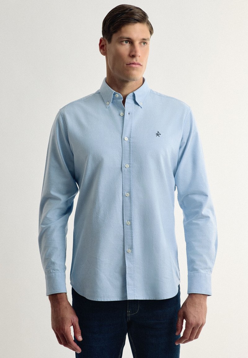 Camisa de botón de manga larga color azul claro con cuello, que presenta un pequeño logo negro en el pecho y una tela suave y texturizada.