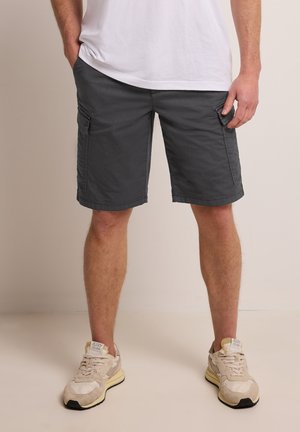 Mann trägt dunkelgraue Cargo-Shorts, weiße Turnschuhe und ein weißes T-Shirt, steht mit den Händen in den Taschen vor einem schlichten Hintergrund.