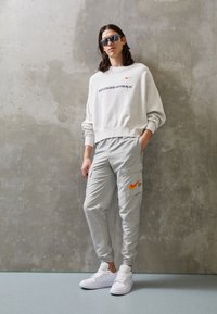Ljusgrå sweatshirt med texten "SWOOSHTRAX" och röd Nike-logotyp, kombinerad med ljusgrå cargobyxor med orange Nike-logotyp och vita sneakers.
