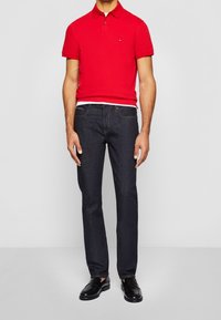 Hombre vestido con una camiseta polo roja, jeans oscuros, mocasines de cuero negros, de pie contra un fondo claro y liso.