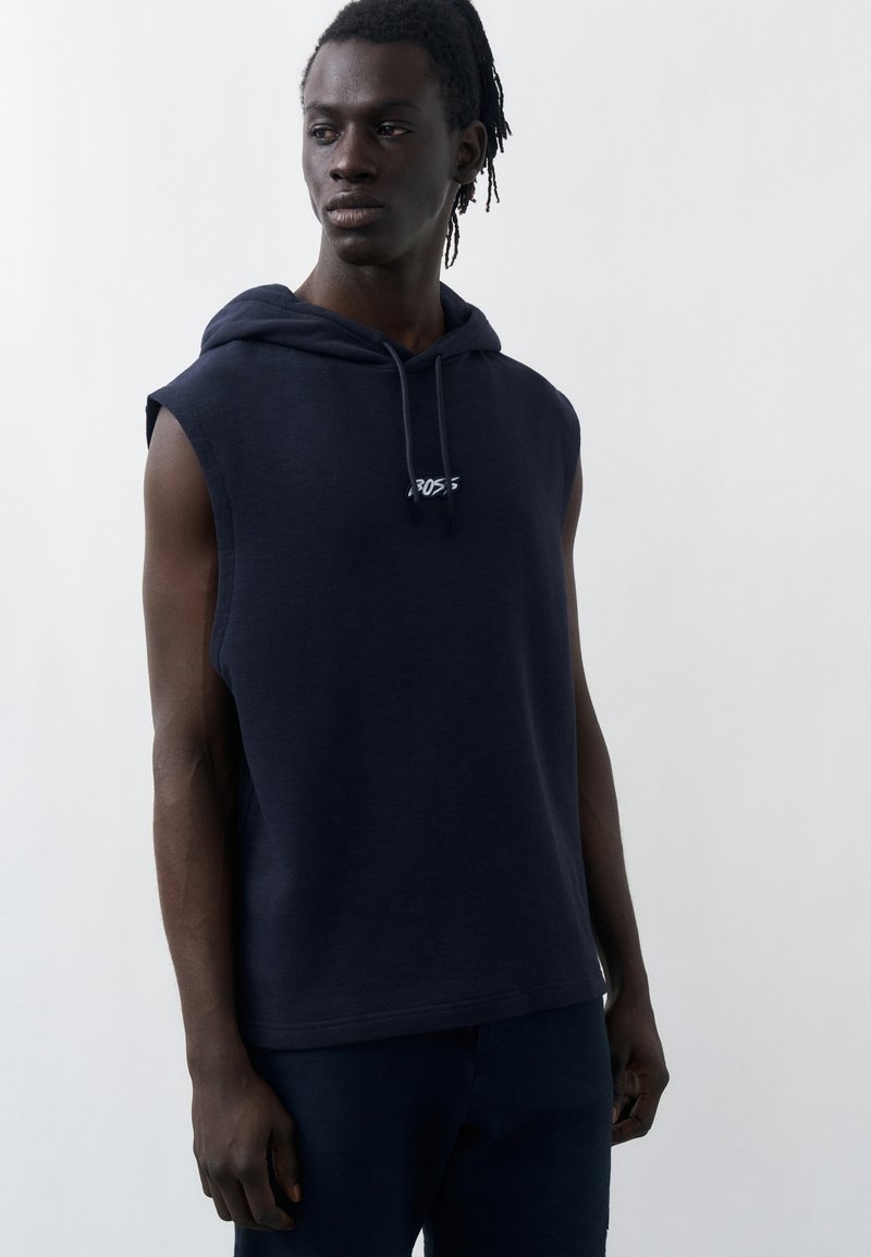 Jeune homme à la peau foncée et aux dreadlocks, portant un sweat à capuche sans manches bleu marine avec un petit logo, debout devant un fond gris uni.