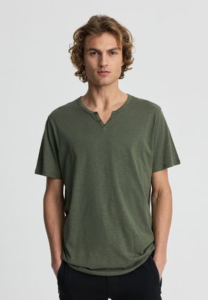 Giovane uomo con capelli biondi ricci, indossa una maglietta henley verde e pantaloni neri, in piedi con le mani in tasca davanti a uno sfondo semplice.