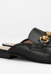 Zwarte leren instappers met een platte hak, voorzien van een gouden metalen accent en een gladde textuur. Ronde neus met minimale stikseldetails.