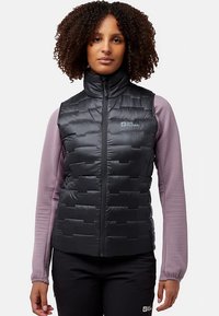 Zwarte puffer-vest met ritssluiting en doorgestikt ontwerp. Gedragen over een roze longsleeve met gestructureerde stof en aansluitende manchetten.