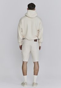 Crème hoodie gecombineerd met lichtbeige denim shorts. Opmerkelijke details zijn de rafelige zoom van de shorts en een leren label op de taille.