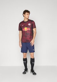 Maillot de football à motifs rouges et noirs avec un logo, associé à un short bleu marine et des chaussettes noires. Chaussures de football noires avec des accents blancs.