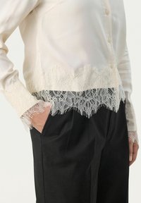 Silke bluse med detaljeret hvid blondestikning ved kanten og manchetterne, med en spids krave og frontlukning med knapper. Parret med sorte bukser.