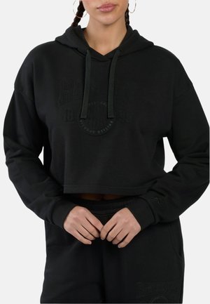 Vrouw draagt zwarte cropped hoodie met trekkoordjes en bijpassende zwarte broek, handen in elkaar gevouwen ter hoogte van de taille, gevlochten haar, grote oorbellen.