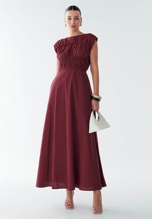 Burgundijas maxi kleita ar platu bodi, īsām kapru piedurknēm un savāktiem vidukļiem; stilizēta ar baltu trīsstūra somu un siksnu papēžiem.