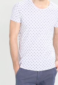 Kortærmet hvid t-shirt med lille blå blomstermønster, rund udskæring og blød bomuldsstruktur, parret med grå bukser.