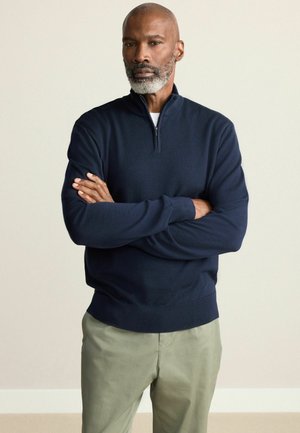 Navy zip-up sweater made of soft knit fabric, featuring a stand collar and ribbed cuffs, paired with light green trousers.
